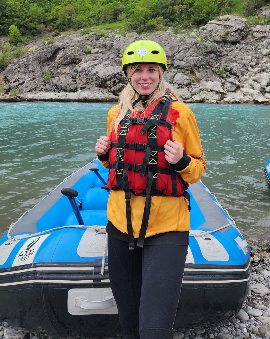 Woman in a helmet and life vest standing by a raft on a river.