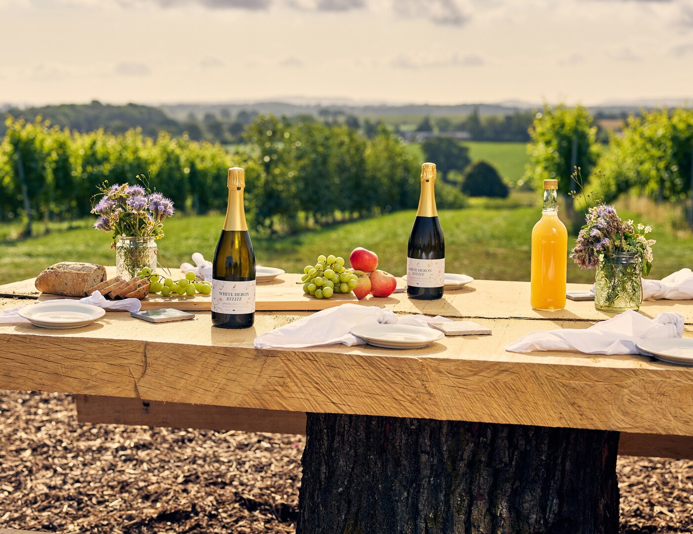 Picnic table with wine, juice, bread, fruit, and flowers, overlooking a vineyard.