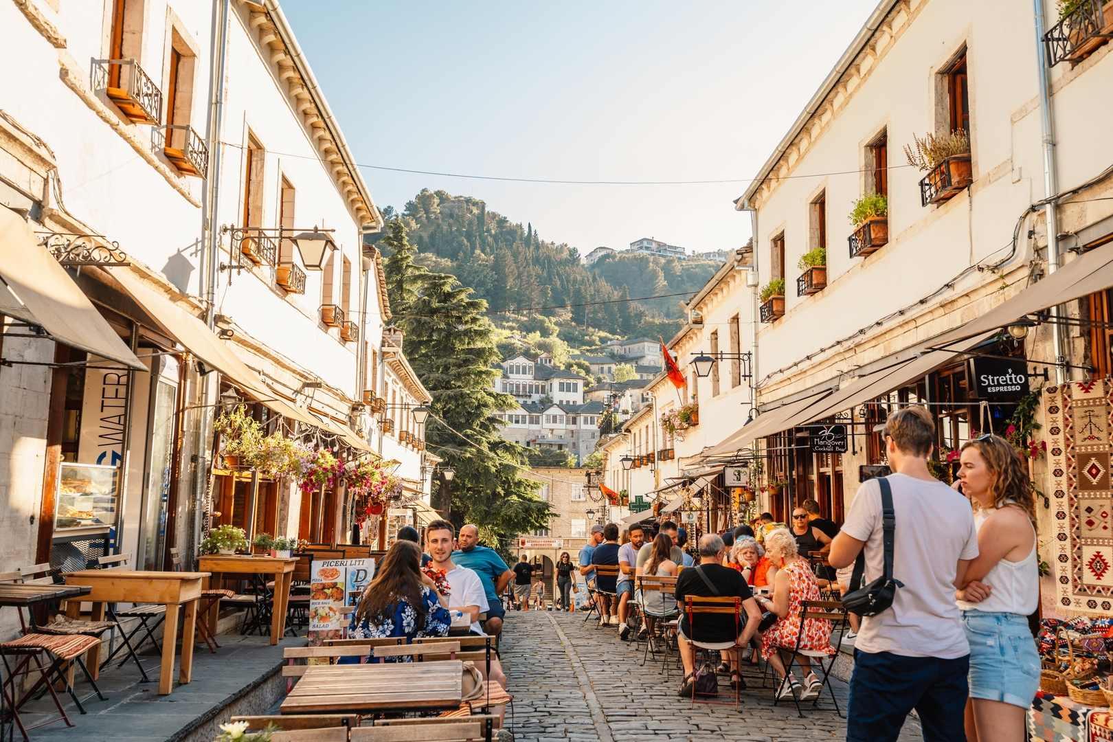 Cobblestone street in Gjirokaster, Albania, lined with outdoor cafes and souvenir shops.
